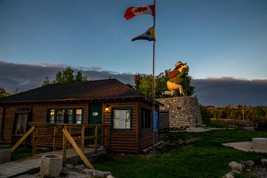 A twilight image of a log building, with a statue of a cartoonish man in old military attire to the right.